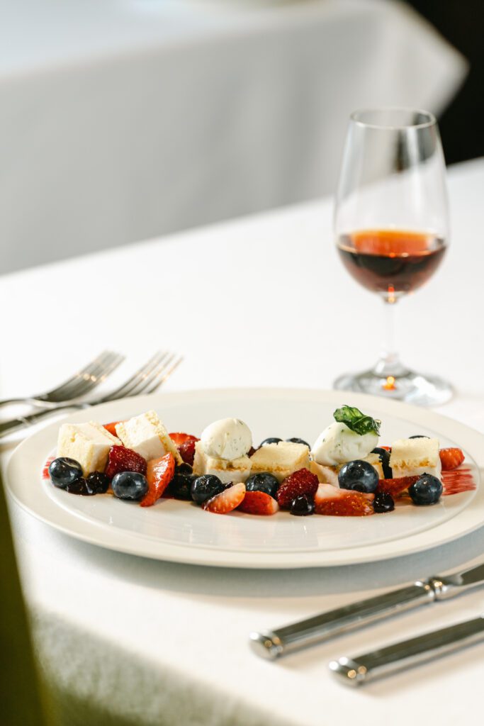 Food and restaurant photography showing a fruit, gelato, and cake dessert with a wine glass of sherry on a white cloth table.
